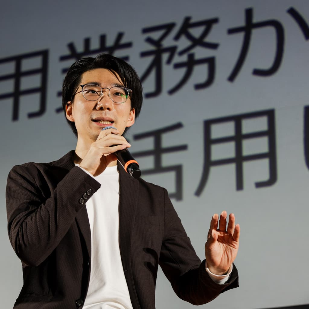 志太悠真 (MacRat) の写真。細い縁のメガネをかけて、白いTシャツに濃いブラウンのジャケットを着ている。巨大なスクリーンの前でハンドマイクを持って、ハンドジェスチャーをまじえながら講演している。